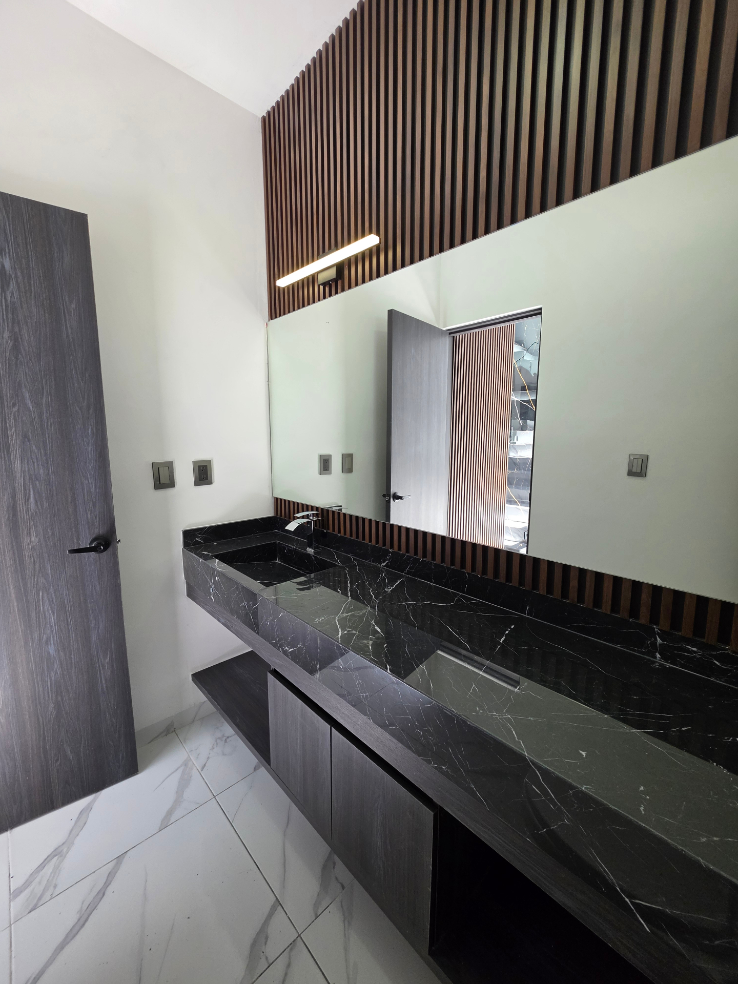 Baño con cubierta de mármol negro, muro de listones de madera y espejo panorámico