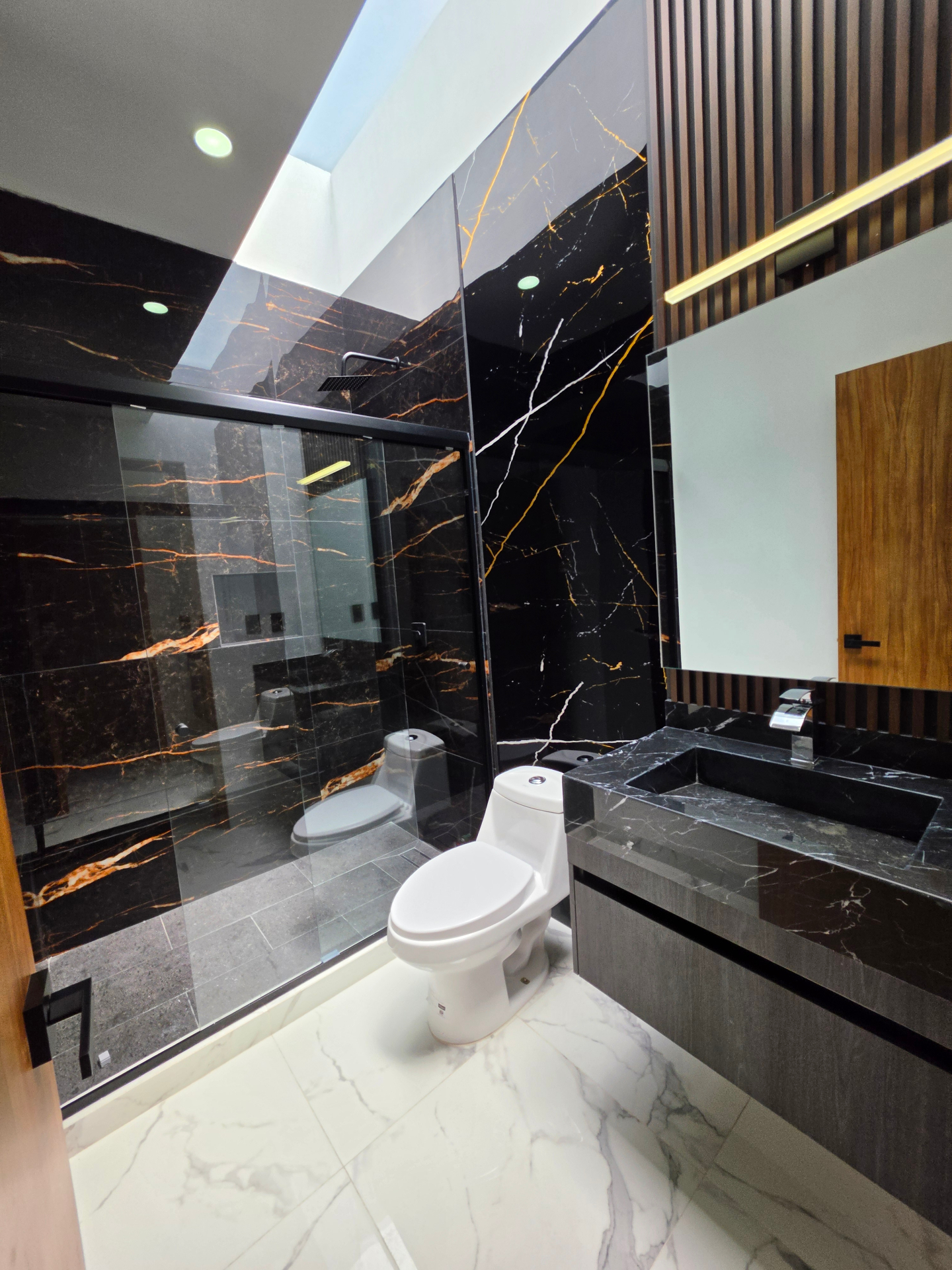 Baño completo con muros negros tipo mármol y cancel de cristal