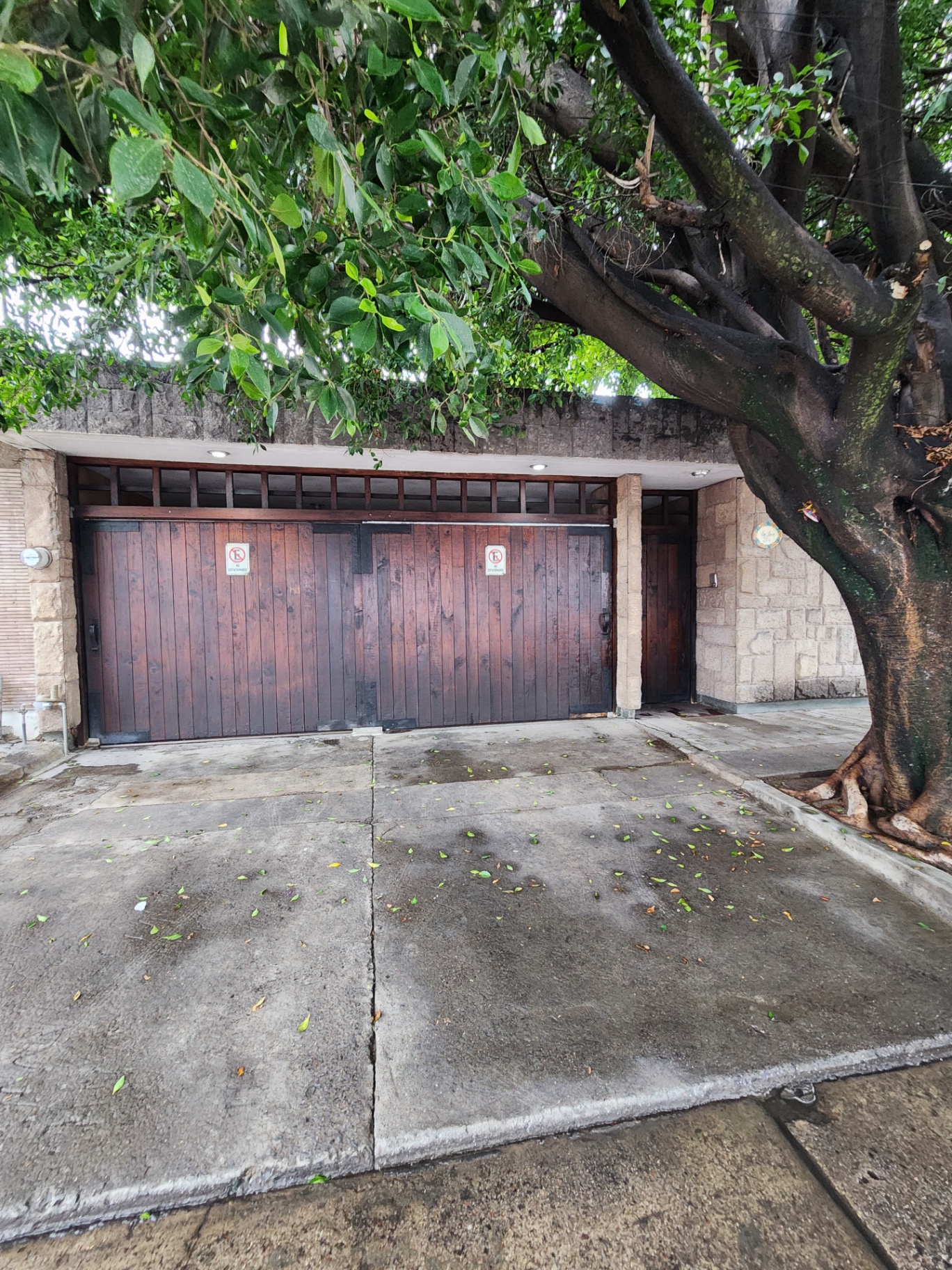 Fachada principal con portón de madera, cochera cerrada para 3 autos y arbolado frontal