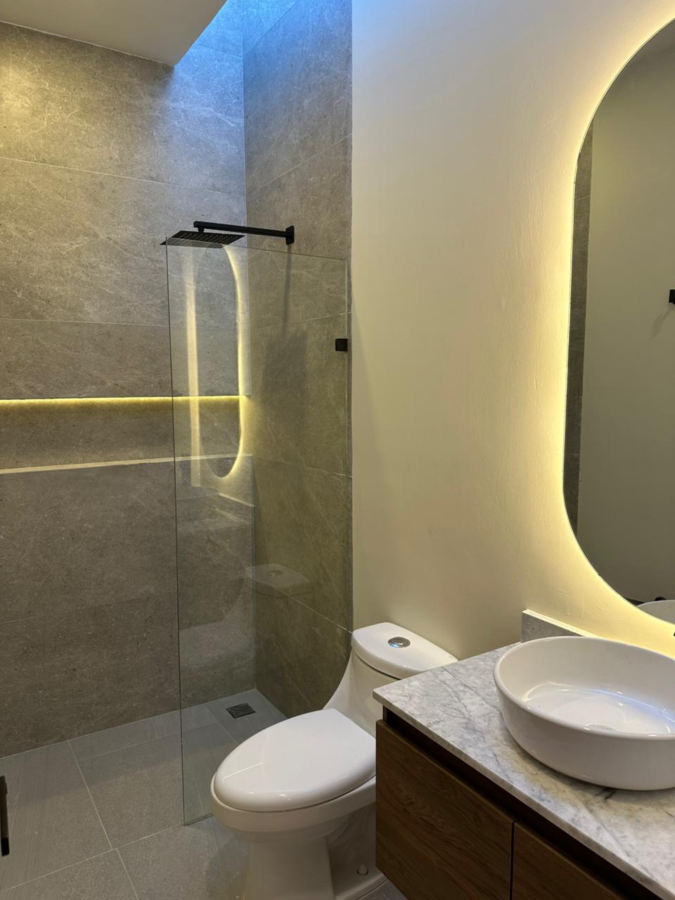 Baño con muro de mármol, regadera de cristal templado, espejo retroiluminado y cubierta de mármol
