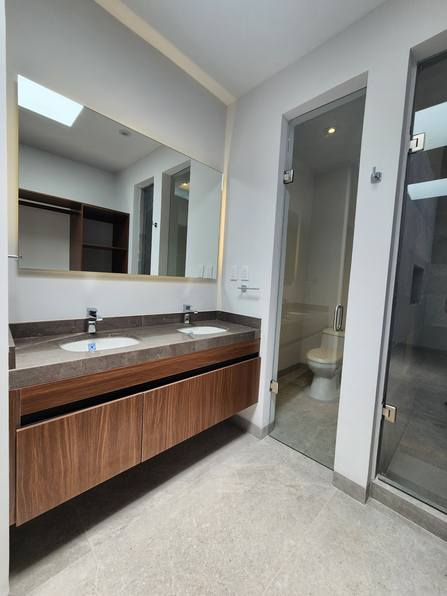 Baño principal con doble lavabo en cubierta de mármol, mueble suspendido en nogal, tragaluz cenital, sanitario en cabina vidriada y regadera independiente