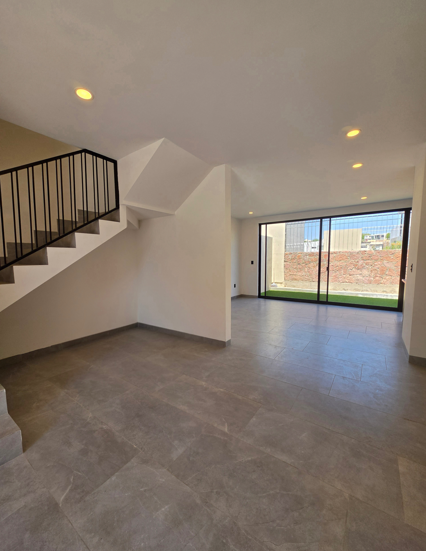 Área social abierta con escalera lateral de barandal negro, piso porcelánico gris y vista al jardín