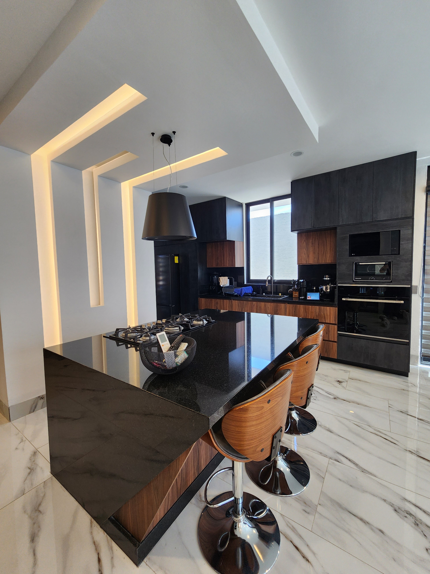 Cocina con isla de granito negro, parrilla integrada, iluminación LED arquitectónica y piso de mármol