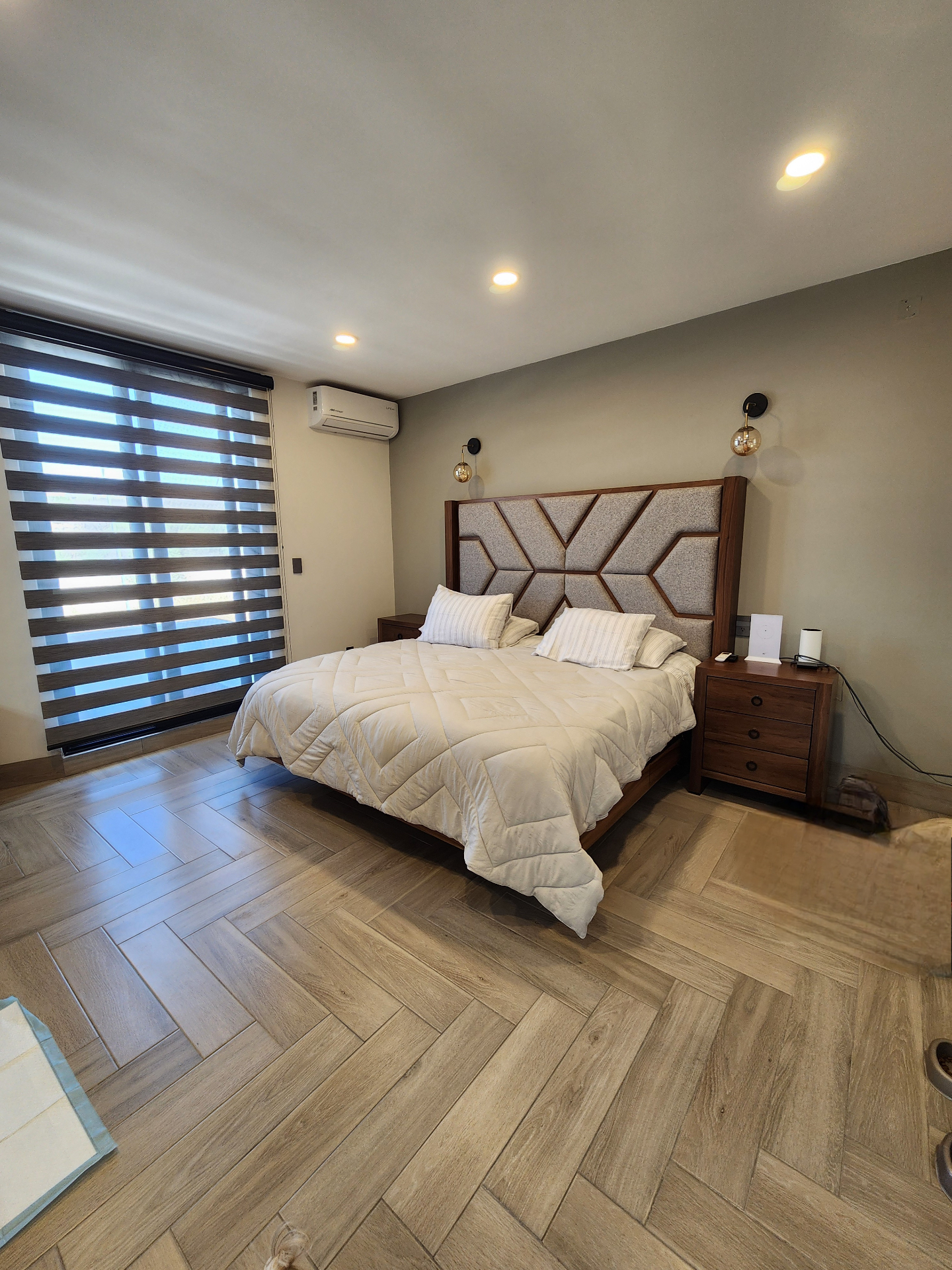 Recámara principal con cabecera de madera y tela geométrica, persianas duo y piso de madera en espiga